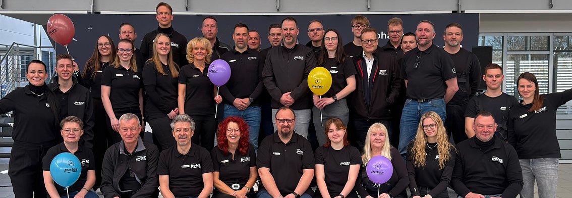 Man sieht ein Teamfoto mit vielen Menschen auf und vor einer kleinen B&uuml;hne. Alle haben das gleiche T-Shirt an, manche halten bunte Luftballons hoch.
