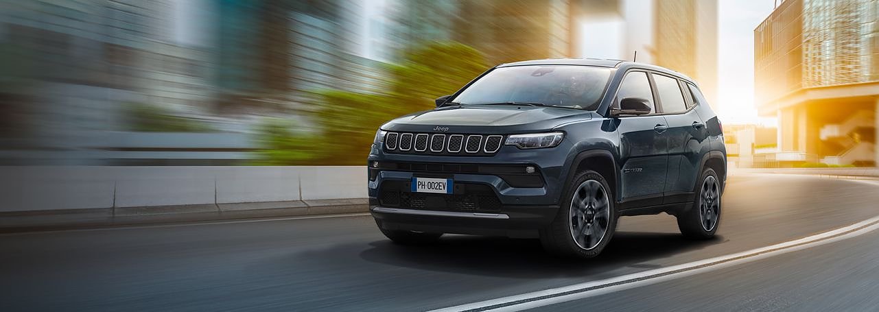 Dunkelgrauer Jeep Compass fährt dynamisch durch eine moderne Stadt mit Hochhäusern und warmem Sonnenlicht im Hintergrund.
