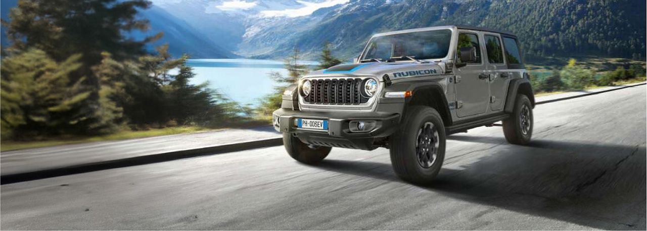 Grauer Jeep Wrangler Rubicon fährt auf einer Straße an einem See vorbei, umgeben von Bergen und Wäldern im Hintergrund.