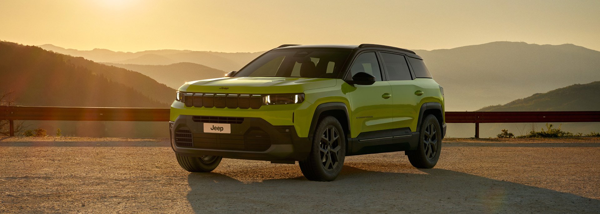 Der neue Jeep® Compass e-Hybrid - Grüner Jeep Compass SUV steht bei Sonnenuntergang auf einem Aussichtspunkt mit Berglandschaft im Hintergrund.
