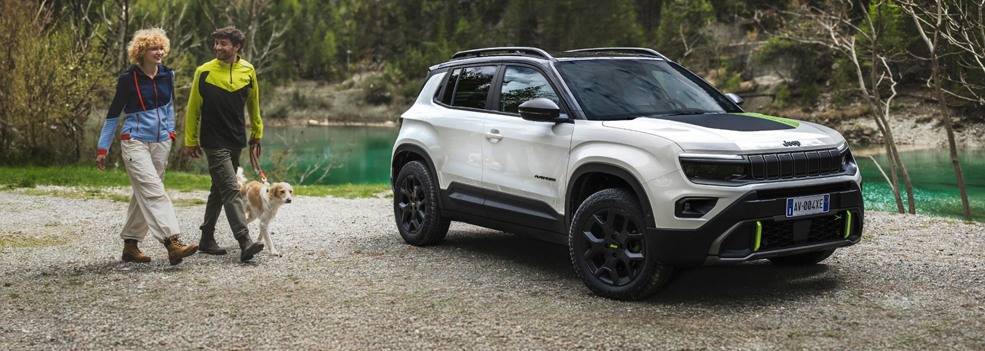 Jeep® Avenger 4xe - Weißer Jeep Avenger 4xe steht vor einem See, daneben gehen zwei Menschen in Outdoor-Kleidung mit einem Hund an der Leine spazieren.