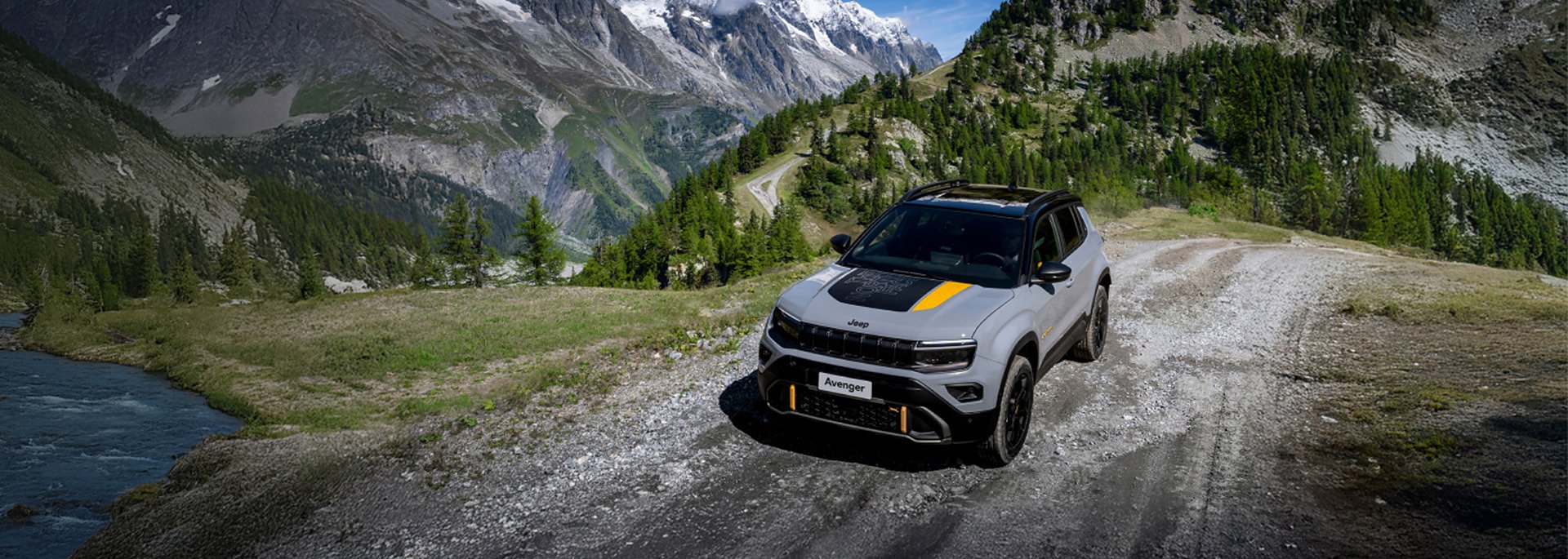 Grauer Jeep Avenger fährt auf einem Schotterweg durch eine alpine Berglandschaft mit Fluss, Wäldern und hohen Felsen im Hintergrund.