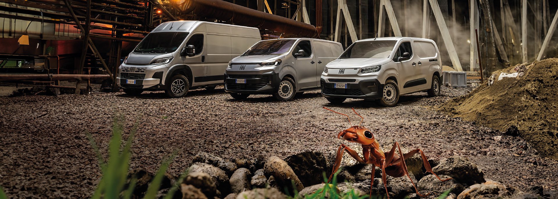 Drei silberfarbene Fiat-Transporter unterschiedlicher Größe stehen nebeneinander auf einem Industriegelände mit grobem Schotterboden. Im Vordergrund ist eine stark vergrößerte rote Ameise zu sehen, die den Eindruck erweckt, sie sei überdimensional groß. Im Hintergrund befinden sich industrielle Rohre und Metallstrukturen mit leichtem Dampf, die eine Baustellen- oder Werksatmosphäre erzeugen.