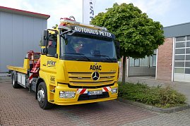 Gelber Abschlepp-LKW des Autohaus Peter im Auftrag des ADAC, Mercedes-Benz, mit der Aufschrift „Autohaus Peter“ auf dem Dachschild und ADAC-Logo an der Front. Das Fahrzeug steht auf einem gepflasterten Platz neben einem Baum und einem grauen Gebäude mit rotem Dach. (Foto: Fischer/Autohaus Peter)