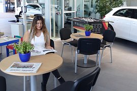 Wartebereich des Duderstädter Mercedes-Autohauses mit einer jungen Frau, die eine Zeitschrift liest, mehreren Tischen mit Zeitungen und Pflanzen, einem Kicker-Tisch im Hintergrund sowie einem weißen Auto, das durch eine Glaswand sichtbar ist. (Foto: Fischer/Autohaus Peter)