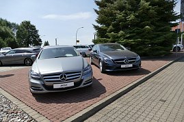 Zwei Mercedes-Benz Fahrzeuge stehen nebeneinander auf einem gepflasterten Parkplatz. Links ein silberner Mercedes und rechts ein dunkler Mercedes. Im Hintergrund weitere Fahrzeuge und große grüne Tannenbäume.  (Foto: Fischer/Autohaus Peter)
