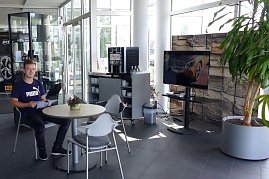Heller Wartebereich im Mercedes-Autohaus mit Glasfront. Ein junger Mann sitzt an einem runden Tisch und liest eine Broschüre. Neben ihm stehen weitere Stühle, ein kleiner Tisch mit einer Pflanze, ein Kaffeeautomat mit Tassenregal und ein großer Fernseher. Rechts im Bild eine große grüne Pflanze in einem grauen Topf. (Foto: Fischer/Autohaus Peter)