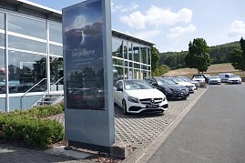 Reihe von Mercedes-Benz Gebrauchtwagen („Junge Sterne“) auf dem Parkplatz eines Autohauses. Im Vordergrund steht ein großes Schild mit dem Logo und der Aufschrift Junge Sterne  Die besten Gebrauchten von Mercedes-Benz. Im Hintergrund moderne Glasfassade des Autohauses, Bäume und Felder bei sonnigem Wetter. (Foto: Fischer/Autohaus Peter)