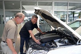 Zwei Männer stehen vor einem weißen Auto mit geöffneter Motorhaube. Einer der Männer trägt ein schwarzes Polohemd und zeigt dem anderen den Motorraum. Beide wirken konzentriert und interessiert. (Foto: Fischer/Autohaus Peter)