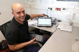 Ein freundlicher Mitarbeiter im schwarzen Polohemd sitzt an einem Schreibtisch in einem Büro. Er hält ein Diagnosegerät in der Hand und zeigt lächelnd auf ein Tablet mit einer Fahrzeug-Software. (Foto: Fischer/Autohaus Peter)