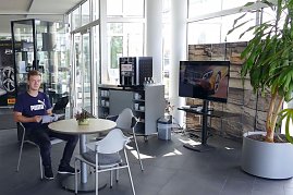 Heller Wartebereich im Mercedes-Autohaus mit Glasfront. Ein junger Mann sitzt an einem runden Tisch und liest eine Broschüre. Neben ihm stehen weitere Stühle, ein kleiner Tisch mit einer Pflanze, ein Kaffeeautomat mit Tassenregal und ein großer Fernseher. Rechts im Bild eine große grüne Pflanze in einem grauen Topf. (Foto: Fischer/Autohaus Peter)