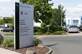 Großes weißes Schild auf dem Gelände eines Mercedes-Benz Autohauses. Auf dem Schild stehen Logos von Mercedes-Benz und Smart sowie Hinweise auf angebotene Leistungen: Verkauf, Service, OMNIplus, Teile/Zubehör, Gebrauchtwagen, Junge Sterne und Van ProCenter. Im Hintergrund sieht man geparkte Autos, grüne Sträucher und Gebäude. (Foto: Fischer/Autohaus Peter)