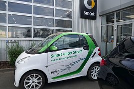 Weiß-grüner Smart mit der Aufschrift Smart unter Strom und die Zukunft gehört DIR!, geparkt vor einem Autohaus. Auf dem Auto ist ein Blitzmotiv abgebildet. Im Hintergrund ist das Smart-Logo an der Gebäudewand zu sehen. (Foto: Fischer/Autohaus Peter)