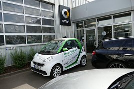 Weiß-grüner Smart mit der Aufschrift Smart unter Strom und die Zukunft gehört DIR!, geparkt vor einem Autohaus. Auf dem Auto ist ein Blitzmotiv abgebildet. Im Hintergrund ist das Smart-Logo an der Gebäudewand zu sehen. (Foto: Fischer/Autohaus Peter)