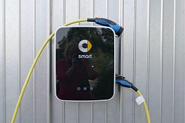 Ladestation mit Smart-Logo an einer silberfarbenen Metallwand. Die Station hat ein schwarzes Frontpanel mit gelbem Kabel und blau-schwarzem Stecker, der an der Seite hängt. (Foto: Fischer/Autohaus Peter)