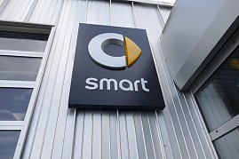 Nahaufnahme eines schwarzen Schildes mit dem Smart-Logo an einer silberfarbenen Metallfassade. Das Logo besteht aus einem stilisierten grauen C mit einem gelben, pfeilförmigen Element, darunter steht der Schriftzug smart in weißen Buchstaben. (Foto: Fischer/Autohaus Peter)