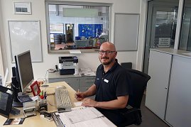 Freundlich lächelnder Mitarbeiter mit Brille und kurzärmeligem Poloshirt sitzt an einem Schreibtisch in einem Autohaus-Büro. Vor ihm liegen Dokumente, ein Computer, Tastatur und Büromaterial. Im Hintergrund ist durch ein Fenster eine Werkstatt mit Reifenstapeln und Werkzeugen sichtbar. (Foto: Fischer/Autohaus Peter)