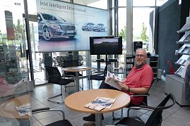 Heller Wartebereich in einem Mercedes-Benz-Autohaus. Ein Mann sitzt an einem runden Tisch, liest eine Broschüre und schaut in die Kamera. Neben ihm liegen Magazine auf dem Tisch. Im Hintergrund ein großer Werbebanner mit Mercedes-Autos, ein Fernseher, Stühle, ein Kaffeeautomat und Regale mit Prospekten. (Foto: Fischer/Autohaus Peter)