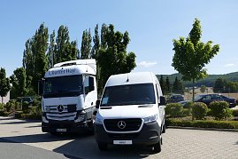 Zwei weiße Mercedes-Benz Nutzfahrzeuge stehen nebeneinander auf einem Parkplatz. Links ein großer CharterWay-Lkw, rechts ein Transporter. Im Hintergrund grüne Bäume, Autos und leicht hügelige Landschaft unter blauem Himmel. (Foto: Fischer/Autohaus Peter)