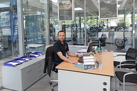 Mitarbeiter in schwarzem Mercedes-Benz Polohemd sitzt an einem modernen Holzschreibtisch im Autohaus, mit Computer, Dokumenten und Stühlen für Kunden, Glaswände mit Werbepostern und Fahrzeugen im Hintergrund. (Foto: Fischer/Autohaus Peter)