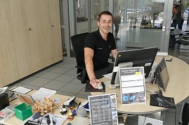 Junger, freundlich lächelnder Mitarbeiter sitzt an einem Schreibtisch im Autohaus. Er trägt ein schwarzes Poloshirt mit Mercedes-Logo. Vor ihm stehen ein Computer, viele Unterlagen, Stempel, Prospekte und Schilder mit Service-Angeboten. Im Hintergrund Glastüren und ein heller Empfangsbereich. (Foto: Fischer/Autohaus Peter)