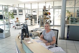 Freundliche Mitarbeiterin sitzt an einem Empfangstresen im Autohaus, vor ihr ein Computer, Telefon und Unterlagen. Sie lächelt in die Kamera. Im Hintergrund sitzt eine weitere Frau an einem Tisch und liest. Helle, moderne Empfangshalle mit Glaswänden, Pflanzen und Schildern, auf einem Glastür-Bereich steht Serviceannahme. (Foto: Fischer/Autohaus Peter)