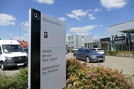 „Schild mit Mercedes-Benz- und Smart-Logo vor einem modernen Autohaus mit Glasfassade. Auf dem Schild stehen Angebote wie Verkauf, Service und Gebrauchtwagen. Davor parken ein grauer Mercedes-Pkw und ein weißer Mercedes-Van. Blumenbeet im Vordergrund, blauer Himmel mit wenigen Wolken im Hintergrund. (Foto: Fischer/Autohaus Peter)