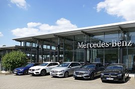 Mercedes-Benz Autohaus mit moderner Glasfassade und großem Mercedes-Benz-Schriftzug. Davor stehen fünf Mercedes-Benz Fahrzeuge in verschiedenen Farben (blau, weiß, silber, grau, schwarz) nebeneinander aufgereiht. Blauer Himmel mit weißen Wolken im Hintergrund. (Foto: Fischer/Autohaus Peter)