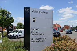 Großes Mercedes-Benz Schild auf einem Autohaus-Gelände. Auf dem Schild stehen die Angebote: Verkauf, Service, Teile/Zubehör, Gebrauchtwagen, Van ProCenter. Im Hintergrund mehrere Mercedes-Fahrzeuge, ein weiteres Smart-Schild und rote Hausdächer. Blauer Himmel mit einigen Wolken. (Foto: Fischer/Autohaus Peter)