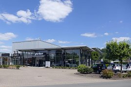 Glasfassade des Autohauses Peter mit Mercedes-Benz und Smart Logos. Vor dem Gebäude stehen mehrere Autos, im Vordergrund Bäume und Büsche. Der Himmel ist blau mit vereinzelten Wolken. (Foto: Fischer/Autohaus Peter)