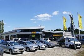Außenansicht des Opel-Autohauses in Osterode mit großem Vorplatz. Im Vordergrund stehen mehrere silberfarbene Opel-Pkw und ein weißer Opel-Transporter mit Kipper-Aufbau. Vor dem Gebäude wehen gelbe und schwarze Opel-Flaggen. Das Autohaus hat eine moderne Glasfassade, darüber sind Schilder mit „Opel Service“ und dem Opel-Logo sichtbar. Der Himmel ist blau mit einigen kleinen Wolken. (Foto: Fischer/Autohaus Peter)
