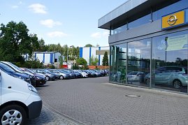 Außenansicht des Opel-Autohauses mit Glasfassade. Rechts ist das gelbe Opel-Logo über dem Eingang sichtbar. Vor dem Gebäude sind viele Autos geparkt, darunter Transporter und verschiedene Pkw-Modelle in einer Reihe. Im Hintergrund sieht man Bäume und Industriegebäude. Der Himmel ist blau mit einigen Wolken. (Foto: Fischer/Autohaus Peter)