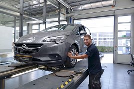 Ein grauer Opel steht auf der Hebebühne der Opel-Werkstatt. Ein junger Mechaniker lächelt in die Kamera, während er mit einem Werkzeug am Vorderrad arbeitet. Im Hintergrund sind große Fenster und ein Garagentor zu sehen, durch das Tageslicht hereinfällt. Auf dem Kennzeichen steht „Opel Zertifizierte Gebrauchtwagen“. (Foto: Fischer/Autohaus Peter)
