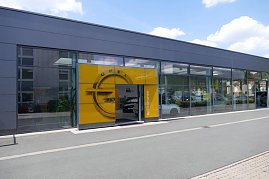 Eingang des Opel-Autohauses Peter in Sondershausen. Die Glasfassade des modernen Gebäudes spiegelt die Umgebung wider, während der gelbe Opel-Eingangsbereich mit großem Markenlogo und „Willkommen“-Schriftzug hervorsticht. Durch die Fenster sind Autos im Innenraum sichtbar. (Foto: Fischer/Autohaus Peter)