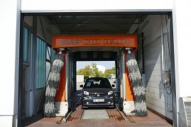Smart-Auto fährt in eine orangefarbene automatische Waschanlage mit rotierenden Bürsten, Schriftzug Christ CAR WASH über dem Eingang. (Foto: Fischer/Autohaus Peter)