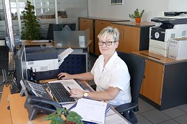 Freundliche Mitarbeiterin sitzt an einem Schreibtisch im Autohaus-Büro, vor einem Computer mit Unterlagen, Drucker und Schränken im Hintergrund (Foto: Fischer/Autohaus Peter)