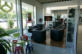 Heller Wartebereich in einem Mercedes-Benz Autohaus mit mehreren schwarzen Ledersesseln, Couchtischen, einem Fernseher und einem Kinderspieltisch mit bunten Stühlen und Spielzeug. Durch die Glasfront ist draußen ein Mercedes-Logo aus Buchsbaum sichtbar. Im Hintergrund Ausstellungsflächen mit Merchandising-Artikeln und ein Kaffeeautomat. (Foto: Fischer/Autohaus Peter)
