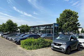 Außenansicht des Mercedes-Benz Autohauses Peter in Dessau. Im Vordergrund steht ein grauer Smart mit dem Schild Gebrauchtwagen. Daneben viele schwarze und silberne Mercedes-Benz Fahrzeuge in einer Reihe auf dem Parkplatz. Im Hintergrund modernes Glasgebäude mit großen Fenstern, Bäumen und blauem Himmel mit leichten Wolken. (Foto: Krüger/Peter-Autozentrum-Anhalt)