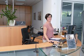 Freundlicher Empfangsbereich im Mercedes-Benz Autohaus in Dessau. Eine Mitarbeiterin hinter einem Holz-Glas-Tresen, hält eine Mappe und lächelt in die Kamera. Auf dem Tresen stehen Stempel, ein Bildschirm, ein Flyerständer und eine Zimmerpflanze. Im Hintergrund Schränke und Bürotüren. (Foto: Fischer/Autohaus Peter)
