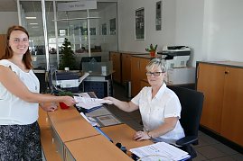 Eine junge Frau überreicht einer Mitarbeiterin an einem Empfangsschalter freundlich ein Dokument. Die Mitarbeiterin sitzt hinter dem Schreibtisch und trägt eine Brille. Beide Frauen lächeln. Im Hintergrund sieht man Büroräume und Schränke. (Foto: Fischer/Autohaus Peter)