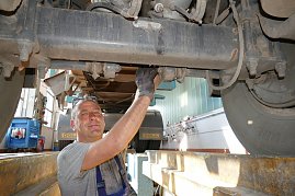 Ein Werkstattmitarbeiter steht unter einem großen LKW und arbeitet an dessen Unterseite. Er trägt graues T-Shirt, schaut in die Kamera und lächelt. Im Hintergrund ist die Werkstatt mit Werkzeugen zu sehen. (Foto: Fischer/Autohaus Peter)
