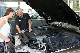 Ein Mercedes-Benz Servicemitarbeiter erklärt einer jungen Frau den Motorraum eines Fahrzeugs. Die Motorhaube ist geöffnet, beide schauen gemeinsam in den Motor. Im Hintergrund helle Glasfronten des Autohauses. (Foto: Fischer/Autohaus Peter)