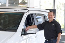Ein Autohaus-Mitarbeiter im schwarzen Hemd mit weißem Mercedes-Logo steht lächelnd neben einem weißen Westfalia-Campervan, lehnt mit einer Hand an der Fahrertür. (Foto: Fischer/Autohaus Peter)