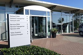 Eingangsbereich des Mercedes-Benz Autohauses in Bernburg mit großen Glasfronten und offen stehender Glastür, davor ein weißes Hinweisschild mit Richtungsangaben zu Verkauf, Service, Zubehör und Gebrauchtwagen, daneben Blumenkübel, gepflasterter Boden und blauer Himmel. (Foto: Fischer/Autohaus Peter)