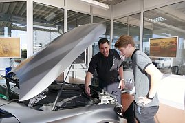Ein Mitarbeiter im schwarzen Hemd mit Mercedes-Logo erklärt einem jungen Kunden Details am Motorraum eines grauen Mercedes-Fahrzeugs. (Foto: Fischer/Autohaus Peter)