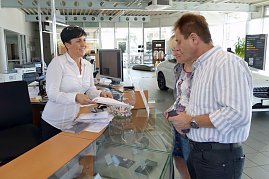 Mitarbeiterin am Empfangstresen im Mercedes-Benz Autohaus Bernburg übergibt lächelnd Unterlagen an ein Paar, moderner heller Showroom mit Computern, Prospekten und Autos im Hintergrund. (Foto: Fischer/Autohaus Peter)