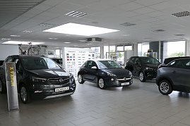 Heller, moderner Showroom des Opel-Autohauses mit mehreren ausgestellten Autos. Links vorne steht ein grauer Opel Mokka X, daneben ein kleinerer Opel Adam in der Mitte. Im Hintergrund sind weitere Fahrzeuge und Fahrräder zu sehen. Die großen Fensterfronten lassen viel Tageslicht herein, die Decke ist mit weißen Paneelen und Einbaulampen ausgestattet. An der Rückwand hängen Farbmuster. (Foto: Fischer/Autohaus Peter)