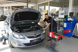 Ein Mechaniker überprüft einen silbernen Opel mit offener Motorhaube in einer Werkstatt. Der Mechaniker steht freundlich lächelnd neben dem Fahrzeug und bedient ein Prüfgerät für die Scheinwerfer. Im Hintergrund sind Schreibtische, Poster und technische Geräte sichtbar. (Foto: Fischer/Autohaus Peter)