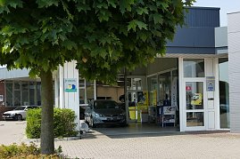 Autohaus-Einfahrt mit einem grauen Opel-Fahrzeug, das unter einem überdachten Bereich steht. Über dem Eingang befinden sich Schilder, unter anderem von DEKRA. Vor dem Gebäude stehen ein Baum mit dichten grünen Blättern und kleinere Büsche. Durch große Glasfronten ist der Innenraum mit Postern sichtbar. (Foto: Fischer/Autohaus Peter)