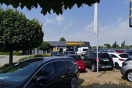 Blick über den Parkplatz eines Opel-Autohauses in Sömmerda. Im Vordergrund stehen viele verschiedene Fahrzeuge, darunter schwarze, rote und weiße Modelle. Im Hintergrund ist das moderne Autohaus mit der markanten gelben Opel-Beschilderung zu sehen, umgeben von Bäumen und Flaggen. Der Himmel ist blau mit ein paar Wolken. (Foto: Fischer/Autohaus Peter)